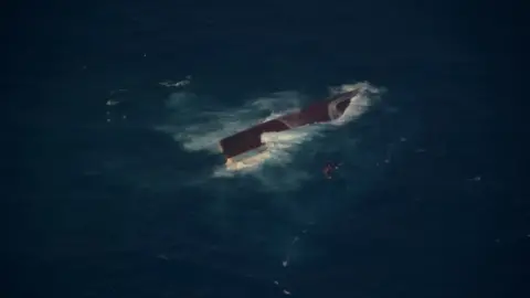 Indonesian Navy Aerial view of a capsized vessel partially submerged in dark blue waters with white foam surrounding the hull