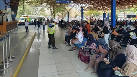 Donny Muslim / BBC People sit and wait inside a busy ferry terminal, while a security guard stands nearby beneath directional signs and an open metal roof