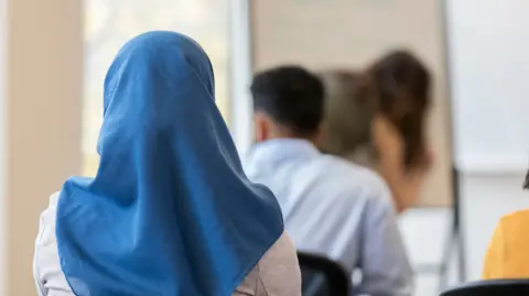 Getty Images A stock image of a girl wearing a blue headscarf. She is sitting down in a classroom, facing with her back towards the camera.